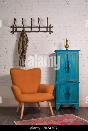 Französischer orangefarbener Ohrensessel mit Holzbeinen, Vintage-blauem Schrank und Wandaufhänger mit kunstvollem Schal an der Wand aus weißen Ziegeln und grunge-Holzparq Stockfoto