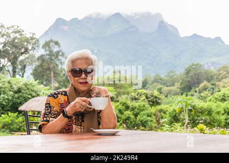 ASIN grau weiße Haare Senior Frau trinken Kaffee im Café im Freien mit Blick auf die Berge. Stockfoto