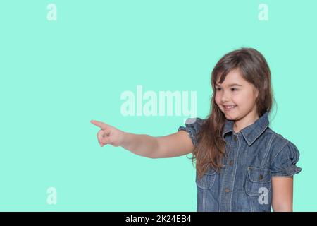 Lächelnd gebräunte langhaarige kleine Mädchen trägt Denim-Kostüm zeigt ihren Finger auf einen leeren Raum bereit für Ihren Text isoliert auf cyanfarbenem Hintergrund. Stockfoto