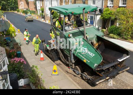 Heißer Tarmakadam, der durch eine asphaltierte Asphaltfertiger-Maschine während der Sanierung einer Wohnstraße in Twickenham, Großraum London, Großbritannien, verteilt wird. Es wird wieder aufgearbeitet, nachdem die zuvor verschlissene und mit Topflappen abgetragene Oberfläche entfernt wurde. (132) Stockfoto