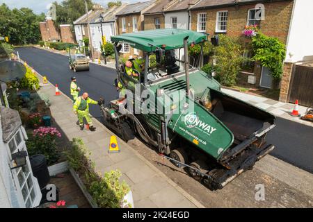 Heißer Tarmakadam, der durch eine asphaltierte Asphaltfertiger-Maschine während der Sanierung einer Wohnstraße in Twickenham, Großraum London, Großbritannien, verteilt wird. Es wird wieder aufgearbeitet, nachdem die zuvor verschlissene und mit Topflappen abgetragene Oberfläche entfernt wurde. (132) Stockfoto