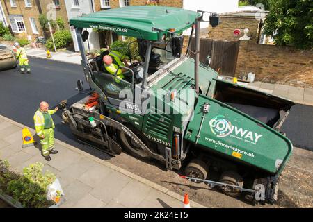 Heißer Tarmakadam, der durch eine asphaltierte Asphaltfertiger-Maschine während der Sanierung einer Wohnstraße in Twickenham, Großraum London, Großbritannien, verteilt wird. Es wird wieder aufgearbeitet, nachdem die zuvor verschlissene und mit Topflappen abgetragene Oberfläche entfernt wurde. (132) Stockfoto