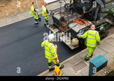 Heißer Tarmakadam, der durch eine asphaltierte Asphaltfertiger-Maschine während der Sanierung einer Wohnstraße in Twickenham, Großraum London, Großbritannien, verteilt wird. Es wird wieder aufgearbeitet, nachdem die zuvor verschlissene und mit Topflappen abgetragene Oberfläche entfernt wurde. (132) Stockfoto