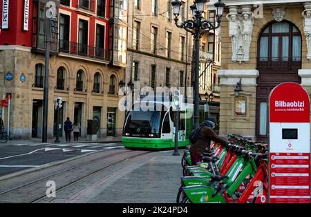 BILBAO, SPANIEN-DECEMBER 19, 2021 : die Stadtbahn Euskotren fährt auf der Straßenbahnlinie auf der öffentlichen Stadtstraße. Menschen an der Bikestation von Bilbaobizi. Nachhaltig Stockfoto