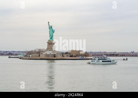 Statue of Liberty National Park, New York City mit weißer ZEPHYR Fähre mit vielen Touristen an Deck vorbei in der Front, während Wintertag wi Stockfoto