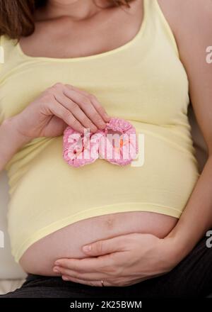 Schwanger mit einer kleinen Prinzessin. Eine junge Schwangerin hält ein Paar rosa Babyschuhe an ihrem Bauch. Stockfoto