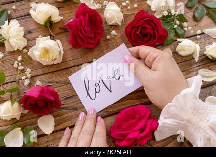 Hände mit Karte LIEBE nahe roten und cremefarbenen Blumen schließen sich auf einem Holztisch Stockfoto