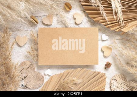 Leere Karte auf einem weißen Holztisch neben getrockneten Pflanzen, Palmenblättern, Pampas-Gras und Herzen Stockfoto