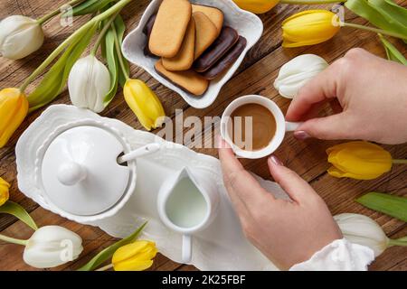 Frauenhände nehmen eine Kaffeetasse aus einem Tablett zwischen weißen und gelben Tulpen auf einem Holztisch Stockfoto