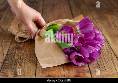 Hand mit lila Tulpenbouquet in Handwerkspapier auf Holztisch Stockfoto