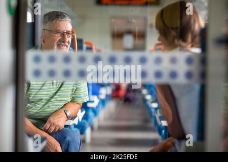 Älterer Mann, der eine Zugfahrt genießt - sein Auto zu Hause lässt, genießt er die Zeit, die er auf Reisen verbringt, und spricht mit anderen Passanten an Bord Stockfoto