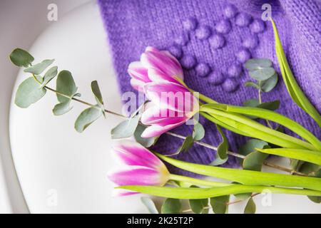 Frühlingsstrauß aus lila Tulpen mit Eukalyptus auf einem weißen Stuhl zusammen mit einem lilafarbenen Pullover. Frühlingsstimmung, 8. März. Postkarte für den Urlaub. Stockfoto