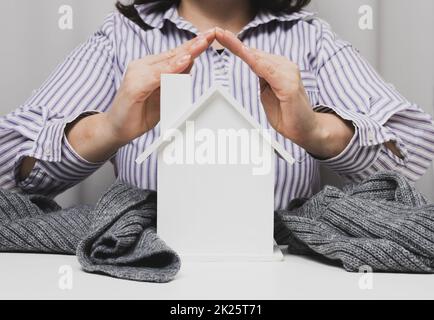 Weibliche Hände zusammengefaltet über einem hölzernen Modellhaus auf weißem Hintergrund. Immobilienversicherungskonzept, Umweltschutz, Familienglück Stockfoto