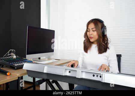 Junge asiatische Frau mit Kopfhörern singen, während sie eine elektrische Tastatur spielt. Musiker, die Musik im professionellen Aufnahmestudio produzieren. Stockfoto