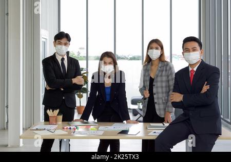 Asiatischer Geschäftsmann in einem Anzug, der sich mit einem selbstbewussten Ausdruck an den Tisch lehnt. Das Team bereitet sich im Hintergrund auf das Meeting vor. Jeder trägt eine Gesichtsmaske. Konzept der Führung. Stockfoto