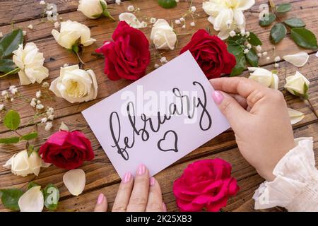 Hände mit Karte FEBRUAR in der Nähe von roten und cremefarbenen Blumen schließen sich auf einem Holztisch Stockfoto