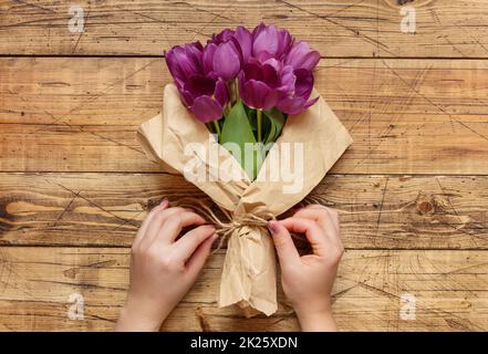 Die Hände wickeln den lilafarbenen Tulpenbouquet in Papier über den Holztisch Stockfoto