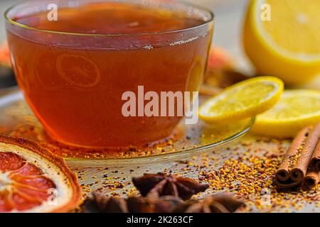 Tasse mit heißem Tee und Dampf auf Schwarz. Glasbecher mit schwarzem Tee mit Zimtstangen, Anisstern, Zitrone und getrockneten Früchten auf weißem Holztisch. Makrobild Stockfoto