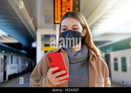 Junge Frau mit schwarzer medizinischer Maske KN95 FFP2 mit Telefon im Bahnhof mit Fahrplänen im Hintergrund Stockfoto