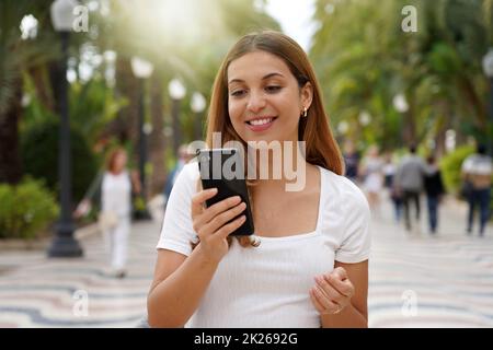 Aufgeregte junge Frau, die ihr Smartphone sieht, wenn sie mit verschwommenen Personen im Hintergrund auf der Straße läuft. Millennials, die im Freien eine mobile App nutzt. Lifestyle-Technologie für Teenager. Stockfoto