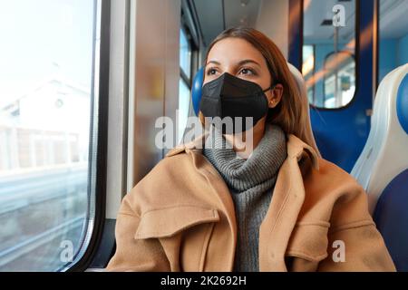 Junge, bedachte Frau trägt schwarze medizinische Gesichtsmaske im Zug und schaut durch das Fenster. Konzept der Reise und Nutzung öffentlicher Verkehrsmittel während einer Pandemie. Stockfoto