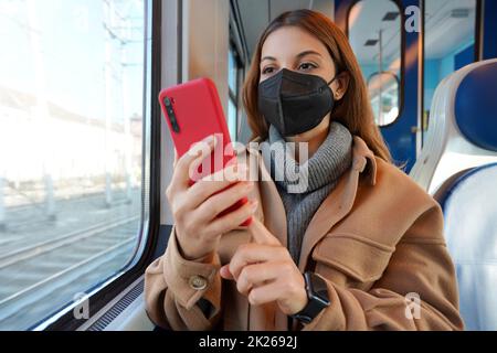 Mädchen in Winterkleidung trägt medizinische schwarze Maske im Zug mit Handy Stockfoto