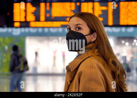 Reisende Frau trägt eine schwarze Schutzmaske KN95 FFP2 am Flughafen, um vor Viren zu schützen. Junge Frau mit Fahrplaninformationen über Abfahrten und Ankünfte auf der Rückseite. Stockfoto