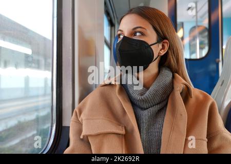 Junge, aufmerksame Frau in einer medizinischen Maske FFP2 KN95 im Zug, die durch das Fenster schaut. Konzept der Reise und Nutzung öffentlicher Verkehrsmittel während einer Pandemie. Stockfoto