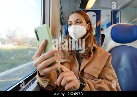 Omicron-Variante. Frau, die mit dem Smartphone im Zug unterwegs ist und eine medizinische Gesichtsmaske trägt. Niedriger Winkel. Stockfoto