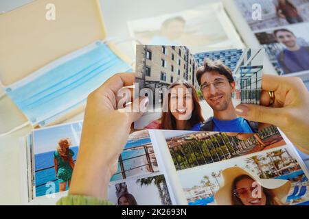 Fernweh während der Pandemie. Draufsicht auf eine Frau, die ihre Bildabzüge mit einer OP-Maske auf einem Taschenkoffer auf dem Hintergrund ansieht. Stockfoto