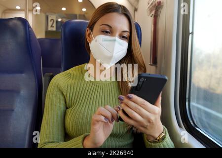 Entspannte Frau im Zug trägt medizinische Gesichtsmaske mit Smartphone-App. Pendler mit Schutzmaske, der in der Business-Klasse sitzt und SMS auf dem Mobiltelefon sendet. Fahren Sie sicher mit öffentlichen Verkehrsmitteln. Stockfoto