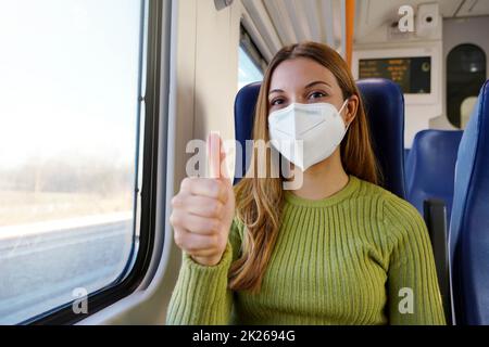 Optimistische junge Frau, die auf öffentlichen Verkehrsmitteln eine schützende medizinische Gesichtsmaske trägt und die Daumen nach oben zeigt, die auf die Kamera schauen Stockfoto