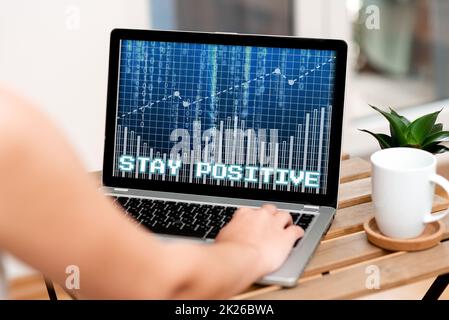 Textanzeige bleibt positiv. Das Wort für „engagieren in aufbauenden Gedanken“ sei optimistisch und tippe neben Coffe Becher und Plant Working From Home auch mit der Hand auf dem Laptop. Stockfoto