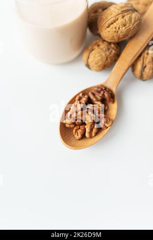 Vegane Milch aus Nüssen in einem Glasbecher mit verschiedenen Nüssen auf einem weißen Tisch, Nüsse in einem Holzlöffel. Stockfoto