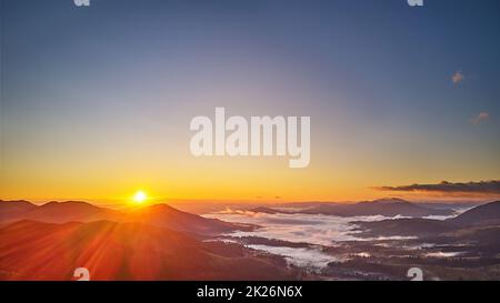 Farbenfroher Sonnenaufgang im Herbst über nebligen Wäldern. Wald bedeckt von niedrigen Wolken. Ländliche Herbstlandschaft. Ein Dorf im nebligen Tal. Stockfoto