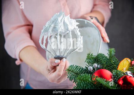 Der Schneebesen eines elektrischen Mischers in einer weißen Masse aus Baiser mit stehenden Gipfeln wird in der Hand eines Konditors gehalten, vor dem Hintergrund einer Eisenkammer, die Zweige eines Weihnachtsbaums und Silvesterspielzeug sind von der Seite sichtbar. Stockfoto