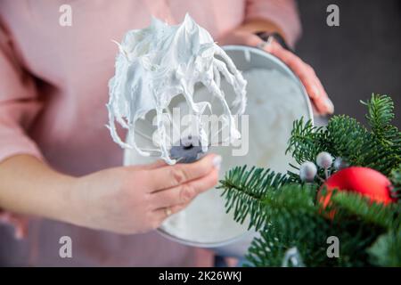 Der Schneebesen eines elektrischen Mischers in einer weißen Masse aus Baiser mit stehenden Gipfeln wird in der Hand eines Konditors gehalten, vor dem Hintergrund einer Eisenkammer, die Zweige eines Weihnachtsbaums und Silvesterspielzeug sind von der Seite sichtbar. Stockfoto
