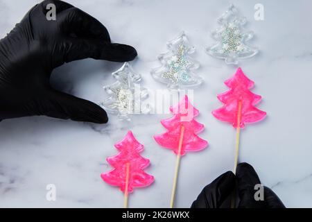 Drei pinkfarbene Lutscher in Form von Weihnachtsbäumen liegen auf weißem Marmor, über ihnen sind weiße Weihnachtsbäume - Lutscher mit Pulver, die extremen halten Hände in schwarzen Handschuhen. Stockfoto