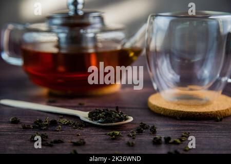 Ein Holzlöffel mit Tee liegt vor einem dunklen Holzhintergrund, ein Becher und eine Teekanne sind von hinten sichtbar, ein Foto in dunklen Farben. Stockfoto