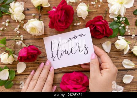 Hände mit KARTENBLUMEN in der Nähe von roten und cremefarbenen Blumen schließen sich auf einem Holztisch Stockfoto