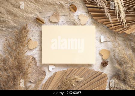 Leere Karte auf weißem Holztisch neben getrockneten Pampas Gras und Herzen, Boho-Modell Stockfoto