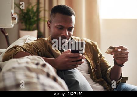 Einkaufen war noch nie so bequem. Freigestellte Aufnahme eines gutaussehenden jungen Mannes, der ein Smartphone und eine Kreditkarte benutzt, um online einzukaufen, während er zu Hause auf seiner Couch sitzt. Stockfoto