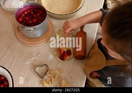 Draufsicht kleiner Bäcker Konditor, der gerollten Teig in Formen mit Kirschen füllt und Tartlets in der heimischen Küche zubereitet Stockfoto