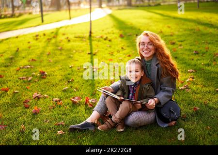 Rotschopf Frau liest Buch mit kleinen Jungen auf Gras im Stadtpark sitzen. Die junge Mutter unterrichtet ihren Sohn mit der ersten Klasse und macht Hausaufgaben am sonnigen Herbsttag Stockfoto