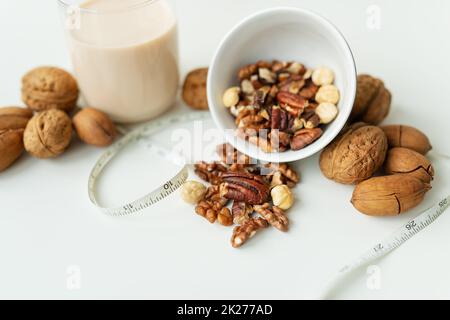 Vegane Milch aus Nüssen in einem Glasbecher mit verschiedenen Nüssen auf einem weißen Tisch. Nüsse in einer Schüssel. Kalorienarme Milch zur Unterstützung des Körpers. Stockfoto