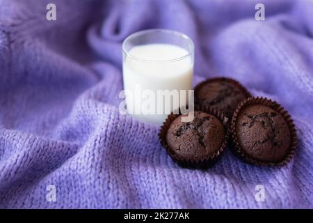 Schokoladen-Cupcakes und ein Glas Milch stehen auf einem lila Strickstoff. Nahaufnahme, Platz für eine Inschrift. Stockfoto