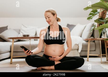 Schwangerschafts- und technologiegestütztes gesundes Lebensstil-Konzept. Fröhliche, glückliche Schwangere, die eine Smartphone-Anwendung verwendet, während sie zu Hause auf der Yoga-Matte im Wohnzimmer trainiert. Stockfoto