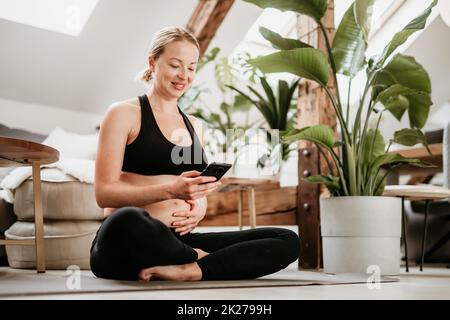 Schwangerschafts- und technologiegestütztes gesundes Lebensstil-Konzept. Fröhliche, glückliche Schwangere, die eine Smartphone-Anwendung verwendet, während sie zu Hause auf der Yoga-Matte im Wohnzimmer trainiert. Stockfoto