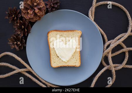 Sandwich zum Frühstück in Herzform mit Käse auf blauem Teller und schwarzem Hintergrund mit Zapfen Stockfoto