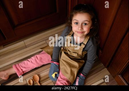 Draufsicht niedliches Kindermädchen, kleine Bäckereikonditorin in beigefarbener Kochschürze und Küchenhandschuhe lächeln süß vor die Kamera Stockfoto